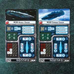 May include: Two Star Wars X-Wing miniatures game cards, one for the MC80 Assault Cruiser and one for the MC80 Command Cruiser. Both cards feature a blue and gray spaceship illustration, a blue and gray ship icon, and a grid of numbers and symbols.