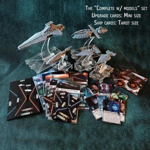 Puede incluir: Una colección de modelos de naves espaciales en miniatura y cartas de juego. Los modelos son grises y están montados sobre soportes transparentes. Las cartas son de varios tamaños y presentan imágenes de personajes y naves. El texto dice "COMPLETE W/ MODELS".