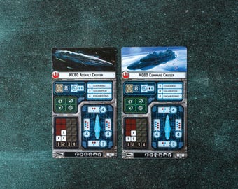 May include: Two rectangular game cards featuring starship illustrations. One card reads "MC80 Assault Cruiser" and the other "MC80 Command Cruiser." Both cards have various game statistics and icons, with a space-themed background.
