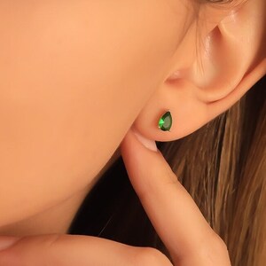 May include: A close-up of a person's ear with a small, teardrop-shaped, emerald green stud earring. The earring is set in a gold or silver metal. The person's hand is touching their neck.
