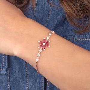 Peut inclure: Un bracelet délicat en or rose avec un pendentif en forme de fleur. Le pendentif est orné de pierres de zircone cubique rouges et claires.