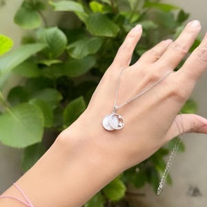 Op de afbeelding: Een zilveren ketting met een witte maan- en sterhanger. De hanger is gemaakt van twee overlappende cirkels, een met een maanontwerp en de andere met een sterontwerp.