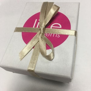 Op de afbeelding: Een witte cadeaudoos met een gouden lint dat in een strik is gebonden. De doos heeft een roze cirkel met de tekst "Live Items" erop.
