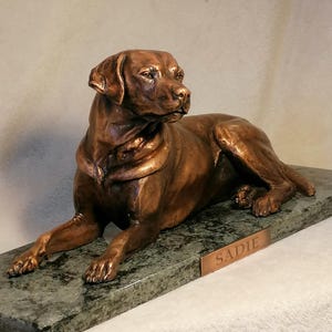 May include: A bronze sculpture of a dog lying down on a green marble base. The dog is a Labrador Retriever and the base has a plaque with the name "SADIE" engraved on it.