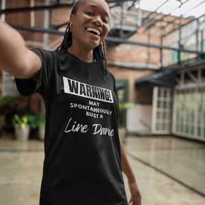 Puede incluir: Una persona lleva una camiseta negra con el texto "WARNING! MAY SPONTANEOUSLY BUST A LINE DANCE" en blanco. La persona sonríe y mira a la cámara.