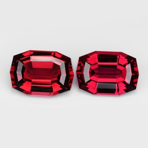 May include: Two faceted, rectangular-cut, red gemstones. The gems have a deep, rich color and are set against a white background. The facets create a play of light and shadow, enhancing their brilliance. These gems are likely for jewelry.