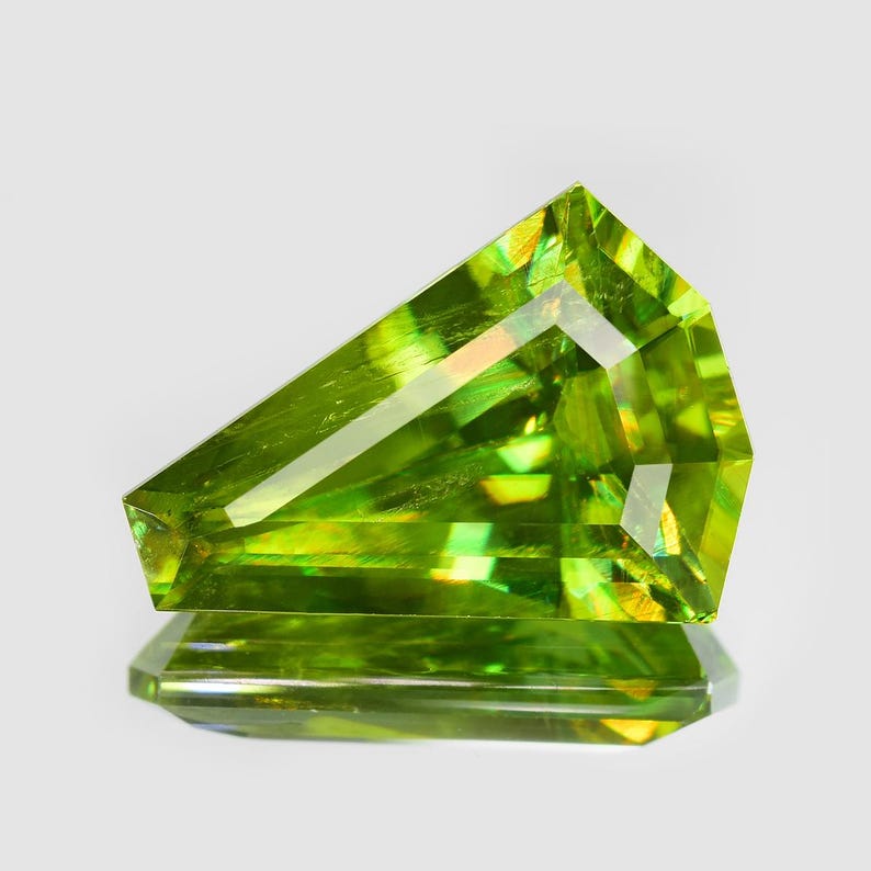 May include: A faceted green gemstone with a unique, asymmetrical cut. The gem has a golden hue and is cut into a sharp, angular shape.