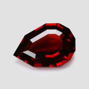 May include: A faceted, pear-shaped gemstone with a deep red hue. The gem has a polished surface with sharp, geometric edges. The color is consistent throughout, with a high degree of clarity, reflecting light. The gemstone is set against a neutral background.