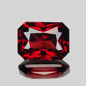 May include: A deep red, rectangular gemstone with a faceted cut, displaying sharp geometric edges and a brilliant, reflective surface. The stone is positioned above its own clear, symmetrical reflection, highlighting its intense crimson color.