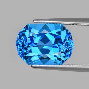 Könnte beinhalten: Ein leuchtend blauer Edelstein mit facettiertem Schliff, gehalten von einer Pinzette. Der Edelstein hat eine rechteckig-ovale Form mit einem brillanten Glanz.