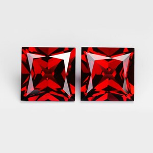May include: Two square-cut, faceted gemstones with a deep red color. The gems have sharp angles and a brilliant, reflective surface, set against a white background. The gemstones are likely for jewelry making or collecting.