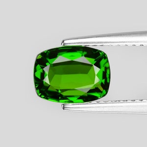 May include: A vibrant green gemstone, likely a peridot, is held by a pair of tweezers. The gemstone is cut into a rectangular shape with rounded edges, showcasing its brilliance and clarity.