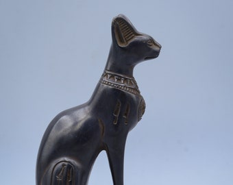 May include: Black Egyptian cat statue with hieroglyphics on the base and body. The cat is sitting with its head turned to the right. The statue is on a small base with hieroglyphics.