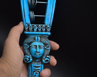 May include: A blue and black wooden sistrum with a carved image of an Egyptian goddess. The sistrum has a handle with hieroglyphics and a frame with metal rings.