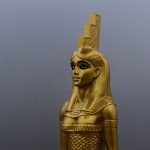 May include: A gold-coloured statue of a woman with long hair and a headdress. The statue features a detailed design, including a scaled dress and armbands. The figure is set against a grey background, highlighting the statue's intricate details and golden colour.