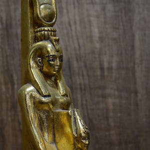 Może przedstawiać: Złota statua egipskiej bogini trzymającej mniejszą statuę faraona. Bogini nosi koronę i długą, falującą szatę. Faraon nosi nakrycie głowy i krótką szatę.