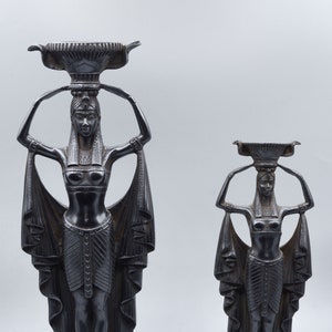 May include: Two black metal statues of Egyptian women holding bowls on their heads. The statues are standing on square bases with hieroglyphics.