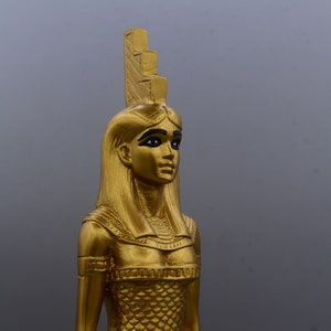 May include: A gold statue of an Egyptian figure, possibly a goddess or pharaoh. The statue has long hair, black eyeliner, and a detailed headdress. The figure is wearing a textured dress and has a serene expression. The statue is approximately 30 cm tall.