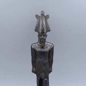 May include: Black statue of an Egyptian pharaoh wearing a tall, pointed headdress. The statue is standing on a rectangular base with hieroglyphics.