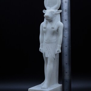 May include: White marble statue of an Egyptian god with a bull's head, wearing a traditional headdress and kilt. The statue is standing on a base with hieroglyphics. The statue is approximately 11 inches tall.