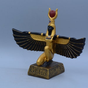 May include: Gold and black statue of the Egyptian goddess Bastet, with outstretched wings and a red jewel on her headdress. The statue is on a square base with hieroglyphics.