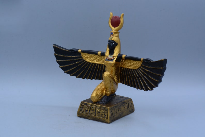 May include: Gold and black statue of the Egyptian goddess Bastet, with outstretched wings and a red jewel on her headdress. The statue is on a square base with hieroglyphics.