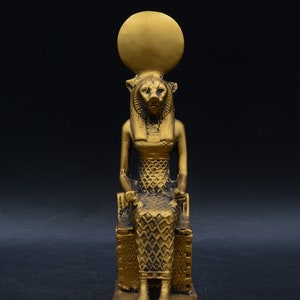 Peut inclure: Statue dor&eacute;e repr&eacute;sentant une figure assise &agrave; t&ecirc;te de lion, v&ecirc;tue d'une robe &agrave; motifs et coiff&eacute;e d'un disque solaire. La statue est sur une base rectangulaire, sur fond sombre.