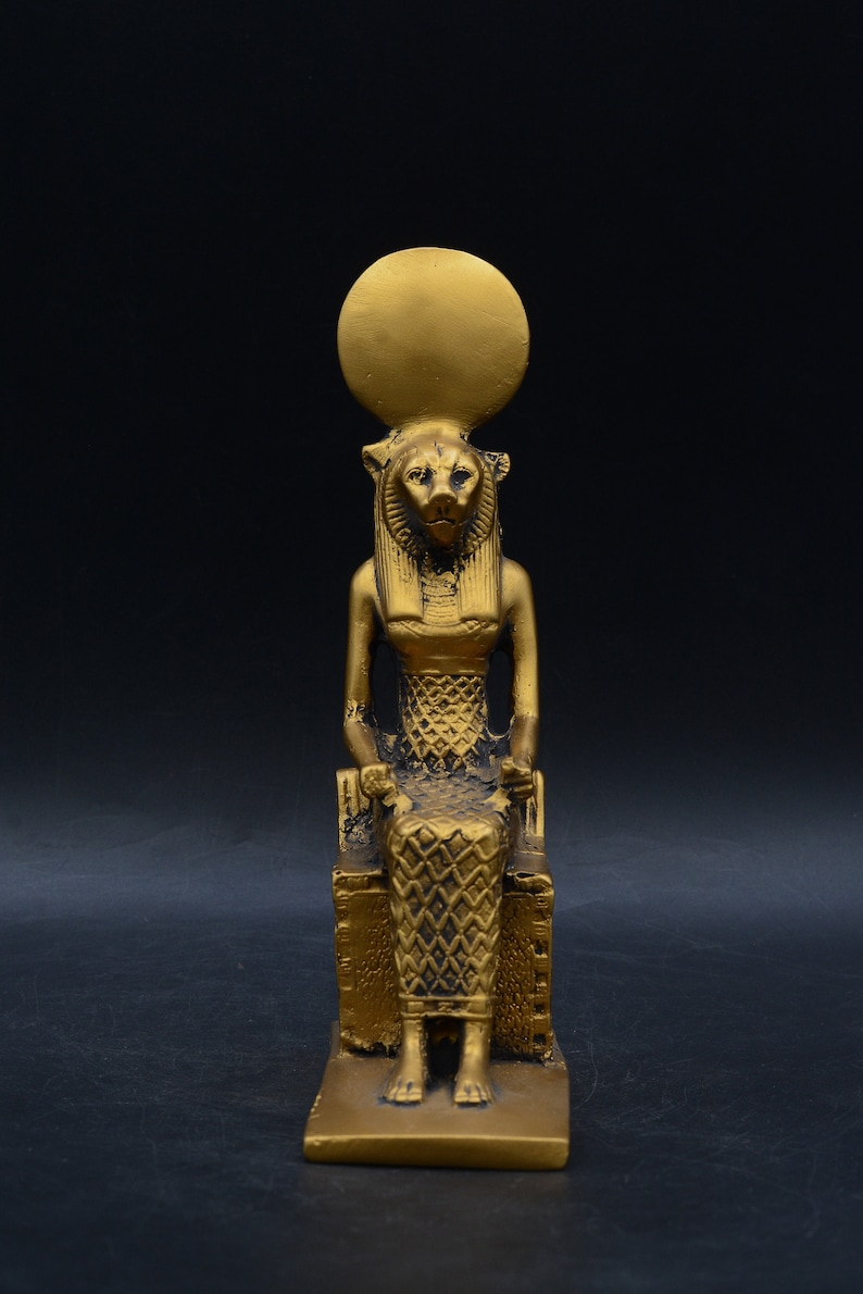 Peut inclure: Statue dor&eacute;e repr&eacute;sentant une figure assise &agrave; t&ecirc;te de lion, v&ecirc;tue d'une robe &agrave; motifs et coiff&eacute;e d'un disque solaire. La statue est sur une base rectangulaire, sur fond sombre.