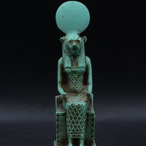 Peut inclure: Statue turquoise d'une divinit&eacute; &eacute;gyptienne, peut-&ecirc;tre Sekhmet, assise sur un tr&ocirc;ne. La figure a une t&ecirc;te de lion, un disque solaire et des d&eacute;tails complexes sur les v&ecirc;tements. La statue est en mat&eacute;riau vieilli, sugg&eacute;rant un aspect antique.