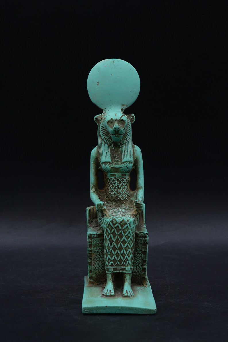 Peut inclure: Statue turquoise d'une divinit&eacute; &eacute;gyptienne, peut-&ecirc;tre Sekhmet, assise sur un tr&ocirc;ne. La figure a une t&ecirc;te de lion, un disque solaire et des d&eacute;tails complexes sur les v&ecirc;tements. La statue est en mat&eacute;riau vieilli, sugg&eacute;rant un aspect antique.