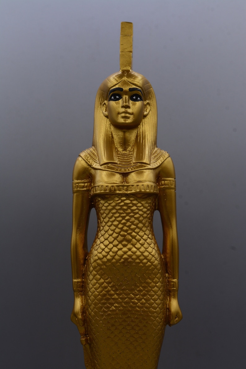 May include: A gold-coloured statue of a woman with a headdress and long hair. The statue has a detailed, scaled dress and is posed with arms at the sides. The figure has black painted eyes and eyebrows.