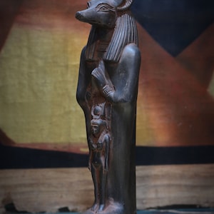 以下が含まれることがあります： ジャッカルの頭をつけたエジプトの神の黒い像で、腕を組んで小さな像を抱えています。像はヒエログリフのある台座の上に立っています。