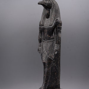 Puede incluir: Estatua de granito negro del dios egipcio Horus, representado como un hombre con cabeza de halc&oacute;n con un disco solar sobre su cabeza. La estatua est&aacute; de pie sobre una base con jerogl&iacute;ficos.