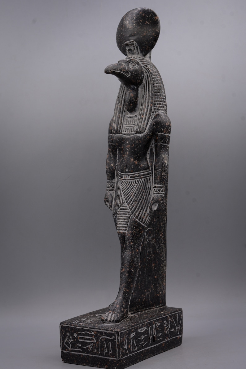 Puede incluir: Estatua de granito negro del dios egipcio Horus, representado como un hombre con cabeza de halc&oacute;n con un disco solar sobre su cabeza. La estatua est&aacute; de pie sobre una base con jerogl&iacute;ficos.