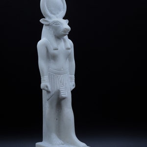 May include: White marble statue of an Egyptian god with bull horns and a human body. The statue is standing on a base with hieroglyphics.