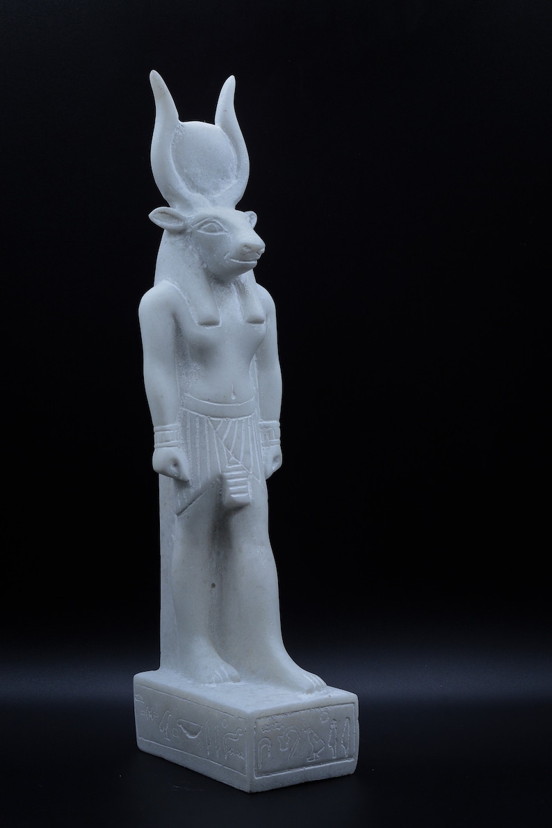 May include: White marble statue of an Egyptian god with bull horns and a human body. The statue is standing on a base with hieroglyphics.