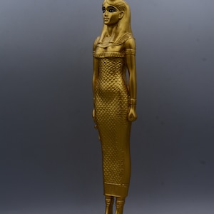 May include: A gold-coloured statue of a woman in a long, scale-patterned dress, standing on a rectangular base. The statue has a detailed headdress and is posed in a standing position. The statue is approximately 30 cm tall.