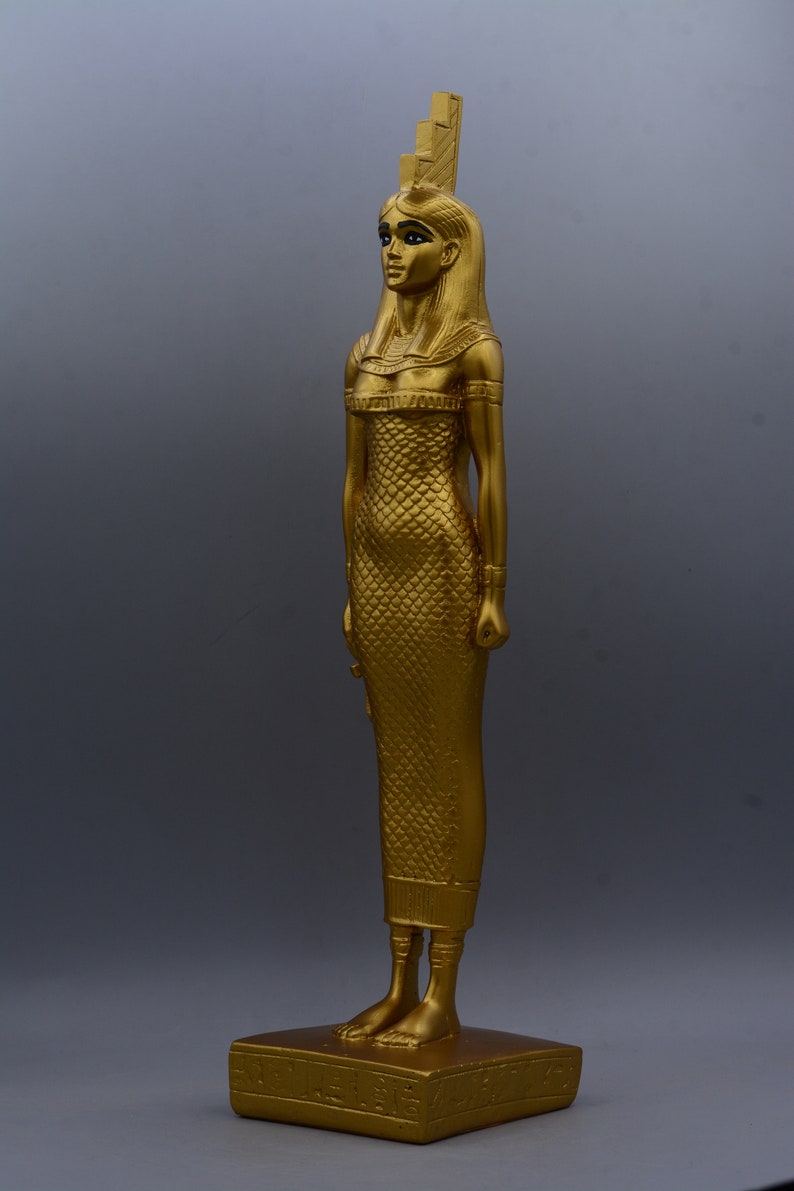 May include: A gold-coloured statue of a woman in a long, scale-patterned dress, standing on a rectangular base. The statue has a detailed headdress and is posed in a standing position. The statue is approximately 30 cm tall.