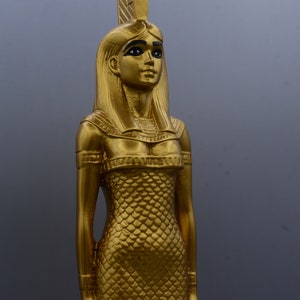 May include: A gold statue of an Egyptian figure, possibly a goddess or pharaoh. The statue has long hair, a headdress, and a detailed dress with a scale-like pattern. The figure is standing and looking forward.