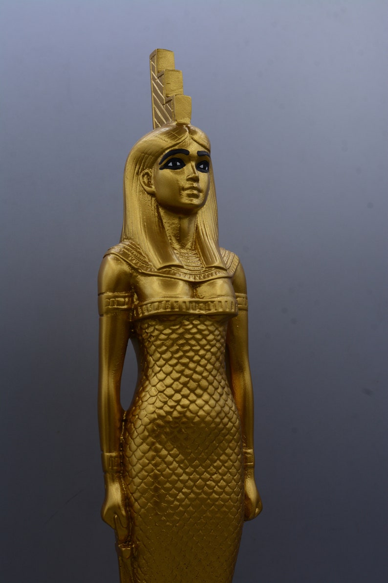 May include: A gold statue of an Egyptian figure, possibly a goddess or pharaoh. The statue has long hair, a headdress, and a detailed dress with a scale-like pattern. The figure is standing and looking forward.