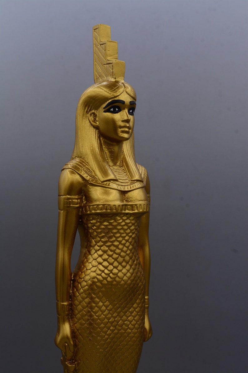 May include: A gold statue of an Egyptian figure, possibly a goddess or pharaoh. The statue has long hair, black eyeliner, and a detailed headdress. The figure is wearing a textured dress and has a serene expression. The statue is approximately 30 cm tall.