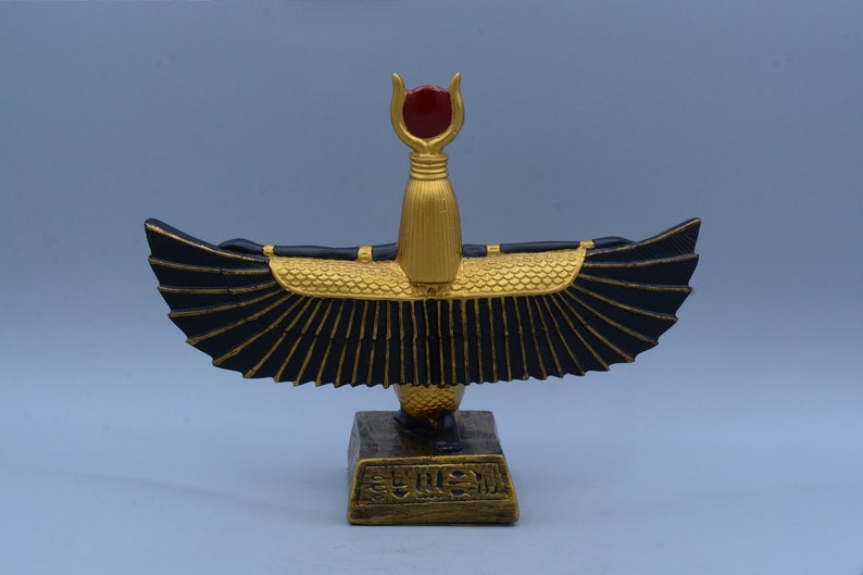 May include: A gold and black statue of the Egyptian goddess Bastet, with outstretched wings and a red gem on her head. The statue is on a base with hieroglyphics.