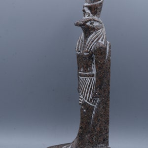 K&ouml;nnte beinhalten: Eine braune Granitstatue des &auml;gyptischen Gottes Horus, dargestellt als falkenk&ouml;pfiger Mann mit einer Krone. Die Statue steht auf einem Sockel mit Hieroglyphen.