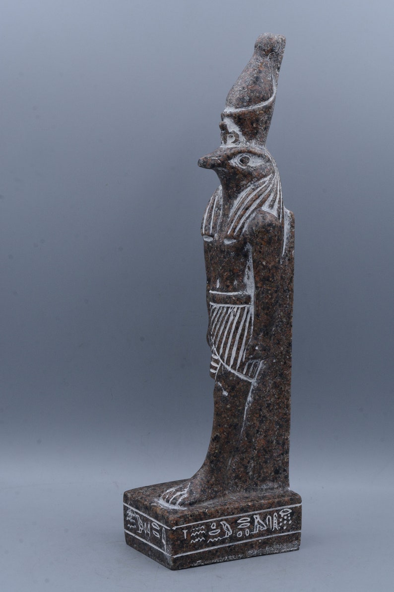 K&ouml;nnte beinhalten: Eine braune Granitstatue des &auml;gyptischen Gottes Horus, dargestellt als falkenk&ouml;pfiger Mann mit einer Krone. Die Statue steht auf einem Sockel mit Hieroglyphen.