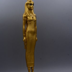 May include: A gold-coloured statue of an Egyptian figure, standing on a rectangular base. The figure wears a long, textured dress and a headdress. The statue is approximately 30 cm tall.