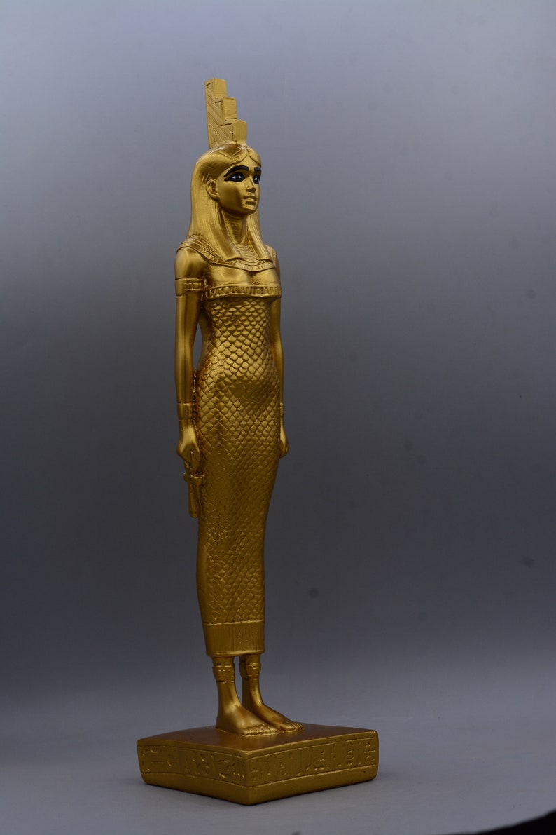 May include: A gold-coloured statue of an Egyptian figure, standing on a rectangular base. The figure wears a long, textured dress and a headdress. The statue is approximately 30 cm tall.