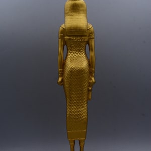 May include: A gold-coloured statue of a figure, possibly a deity, with a tall headdress and a long, patterned dress. The statue stands on a rectangular base. The figure's arms are at its sides, and it has a detailed, textured appearance.