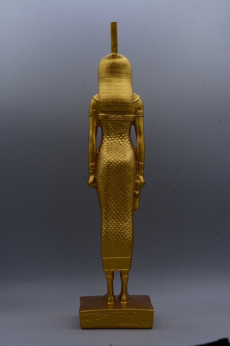 May include: A gold-coloured statue of a figure, possibly a deity, with a tall headdress and a long, patterned dress. The statue stands on a rectangular base. The figure's arms are at its sides, and it has a detailed, textured appearance.