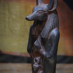 以下が含まれることがあります： ジャッカルの頭をつけたエジプトの神の黒い像。像は腕を胸の前で交差させて立っている。像は複雑なディテールで彫られており、風化したような外観をしている。