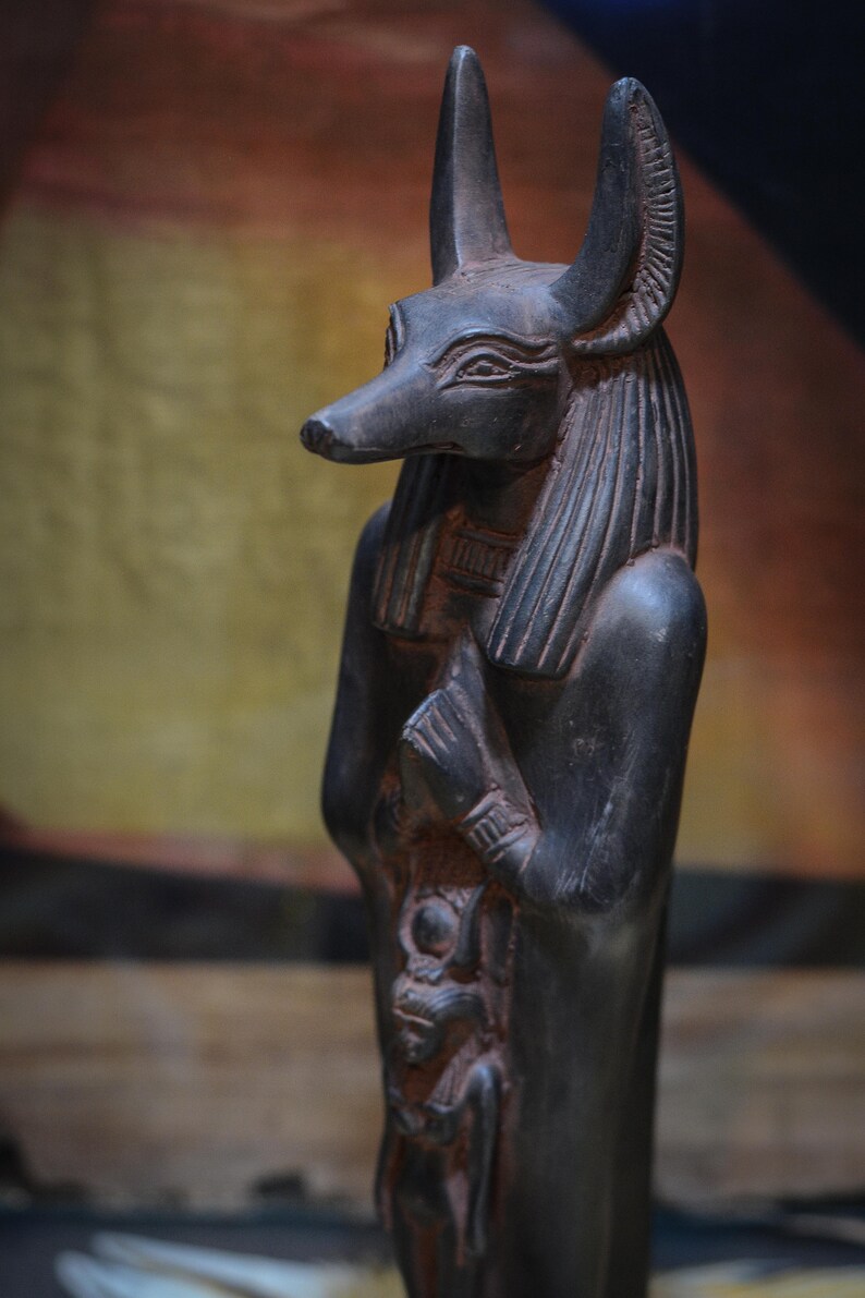 以下が含まれることがあります： ジャッカルの頭をつけたエジプトの神の黒い像。像は腕を胸の前で交差させて立っている。像は複雑なディテールで彫られており、風化したような外観をしている。